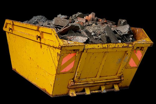 A large, rectangular yellow skip filled with broken construction debris, including chunks of concrete, bricks, and rubble, sits on a black background. The skip has visible metal surfaces with some rust and wear, featuring reflective pink warning markings on its sides. It is positioned on a paved or solid surface, possibly associated with a commercial or industrial waste collection site in Hayes, with no other objects or environmental features visible in the scene. The debris appears freshly dumped and is piled unevenly, indicating recent loading, with some smaller fragments scattered among larger pieces of building material. The lighting highlights the rough textures and varied shades of gray, brown, and reddish tones of the rubble, aligning with typical waste removal processes by companies like Commercial Waste Hayes that handle construction and demolition refuse.