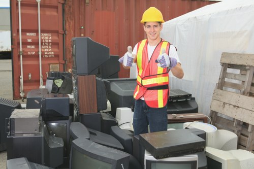 A young male worker in a yellow safety helmet, high-visibility orange vest with reflective stripes, and purple gloves is standing outdoors at a waste disposal site, surrounded by a variety of discarded electronic appliances such as old televisions, computer monitors, and other obsolete equipment. The electronic waste features different sizes and shapes, with visible screens and casing in shades of black, grey, and white. The background includes a rusty shipping container and a stack of wooden pallets, indicating a recycling or disposal environment typical for commercial waste management in Hayes. The worker is smiling and giving a thumbs-up gesture, emphasizing a professional yet approachable attitude, while the cluttered area highlights the importance of responsible rubbish removal services. The setting is well-lit with natural daylight, and the scene reflects a typical scene managed by Commercial Waste Hayes as part of their electronic waste collection and disposal services for businesses in the local area.