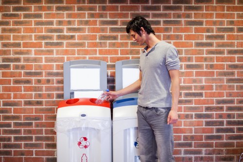 A young man with dark hair, wearing a light grey hoodie and grey jeans, is disposing of a clear plastic bottle into a recycling bin, positioned in front of a brick wall. The recycling bin is white with a red recycling symbol on the front and a red lid, placed beside a similar white bin with a blue lid, both intended for waste separation. The outdoor setting appears to be a paved area, likely part of a commercial or residential environment in Hayes, with the bins lining up against the brick wall. The image conveys waste disposal activity related to rubbish collection services, emphasizing recycling efforts within a local context. Commercial Waste Hayes supplies rubbish removal and waste management solutions, supporting proper environmental practices in the area.